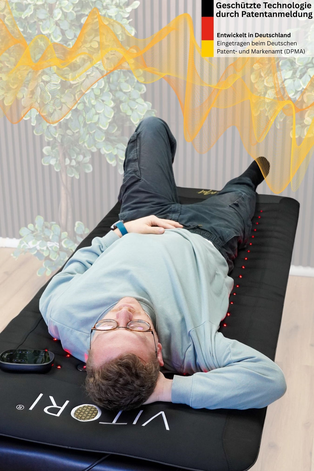 Ein Mann mit Brille entspannt auf einer schwarzen VITORI® VitaMat® mit rotem Licht und VitaWave® Technologie und hält ein Smartphone in der Hand. Oben rechts sind abstrakte gelbe Schallwellen und ein deutscher Patenthinweis zu sehen.