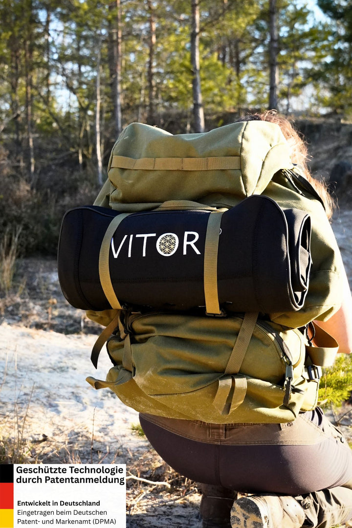 Eine Person mit einem großen grünen Rucksack kniet auf sandigem Boden. Eine schwarze VITORI® VitaMat® mit Infrarot-Tiefenwärme ist am Rucksack befestigt. Sonnenlicht fällt durch die Bäume im Hintergrund.