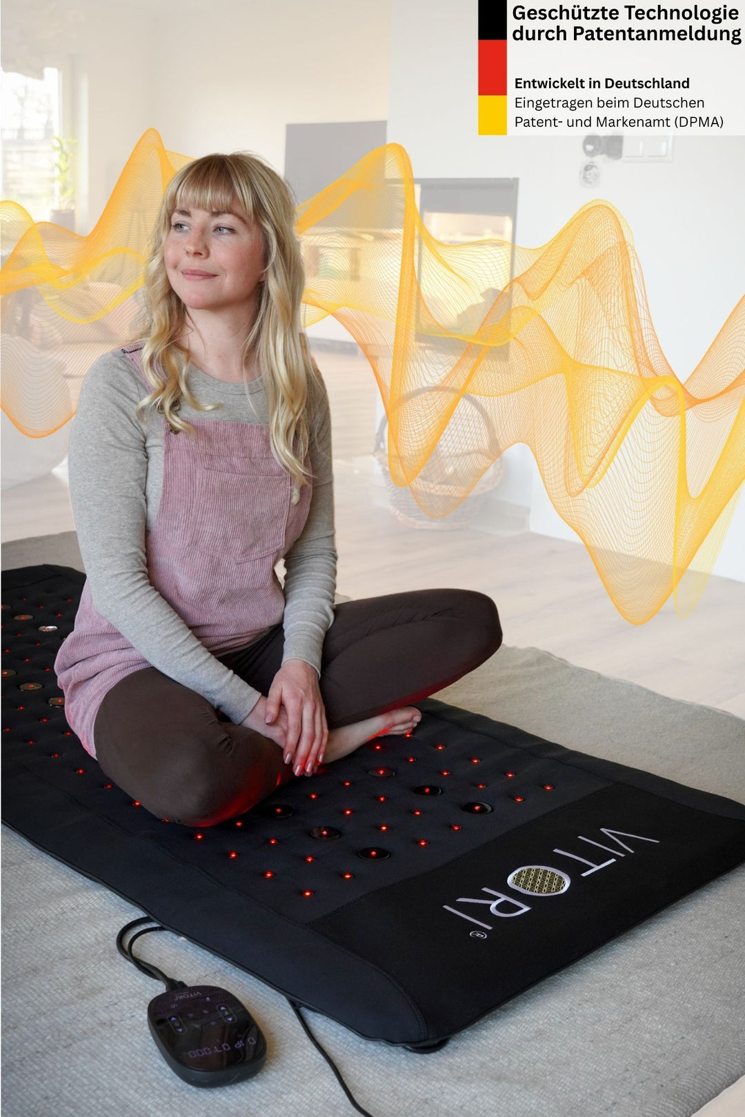 Eine Frau sitzt im Schneidersitz auf einer VITORI® VitaMat®, einer schwarzen Matte mit roten LED-Leuchten, in einem hellen Wohnzimmer. Oben sind gelbe Schallwellengrafiken, ein deutsches Patent sowie Informationen zum VitaWave®-Technologie-Konzept zu sehen. Marke: VITORI.
