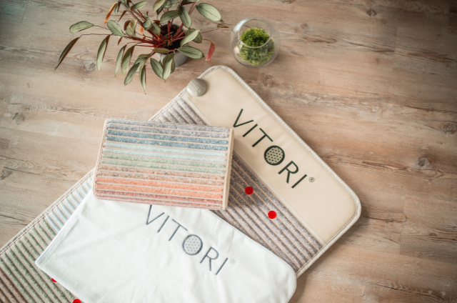 Ein VITORI® Paket Ultimate wird auf einem Holzboden mit einem gestreiften Kissen, einem Kristallkissen, einer Glasschale mit Moos und einer grünen Blattpflanze arrangiert und sorgt so für die ultimative saubere, minimalistische Atmosphäre.