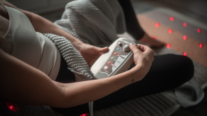 Eine Person sitzt mit einer grauen Strickdecke auf dem Boden und hält einen Controller in der Hand. Daneben befinden sich rote LED-Leuchten und die VITORI® Premium Kristallmatte – Das Original, die auf die Anwendung von Lichttherapie oder Infrarot-Tiefenwärme hindeuten.