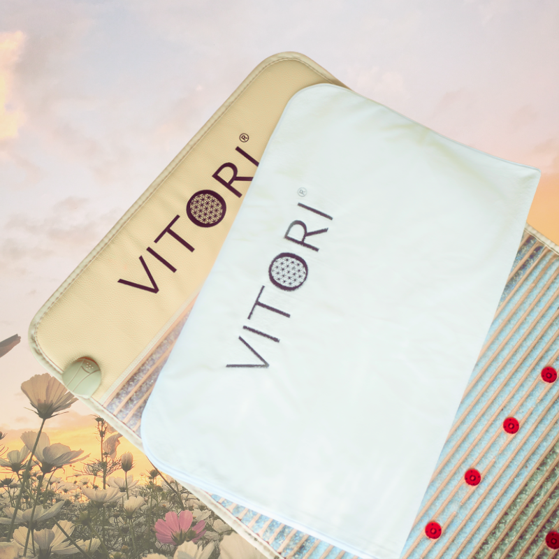 Zwei VITORI® Paket Essential Matten, eine beige und eine weiße, liegen auf einem weichen Hintergrund mit Blumen und einem bewölkten Sonnenuntergang. Rote Knöpfe entlang der beigen Matte vermitteln Ausgeglichenheit und Entspannung in dieser ruhigen VITORI®-Szene.