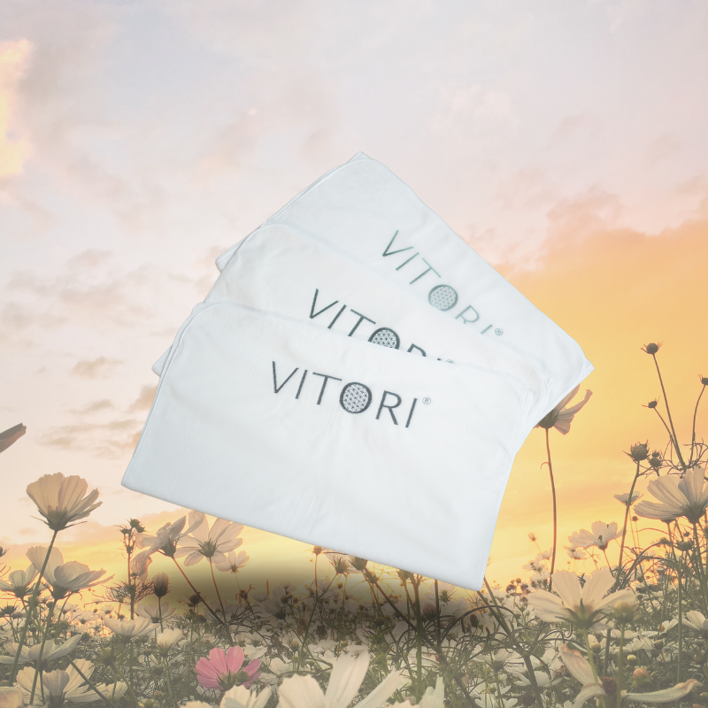 Drei weiße Handtücher mit dem VITORI®-Logo liegen vor einem sanften Sonnenuntergangshimmel. Jedes ist mit dem Bezug für Premium Kristallmatte versehen, die für höchsten Komfort und Hygiene sorgt.