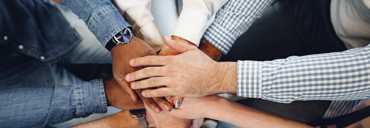 Verschiedene Menschen legen ihre Hände übereinander im Kreis – Symbol für Teamarbeit und Zusammenhalt